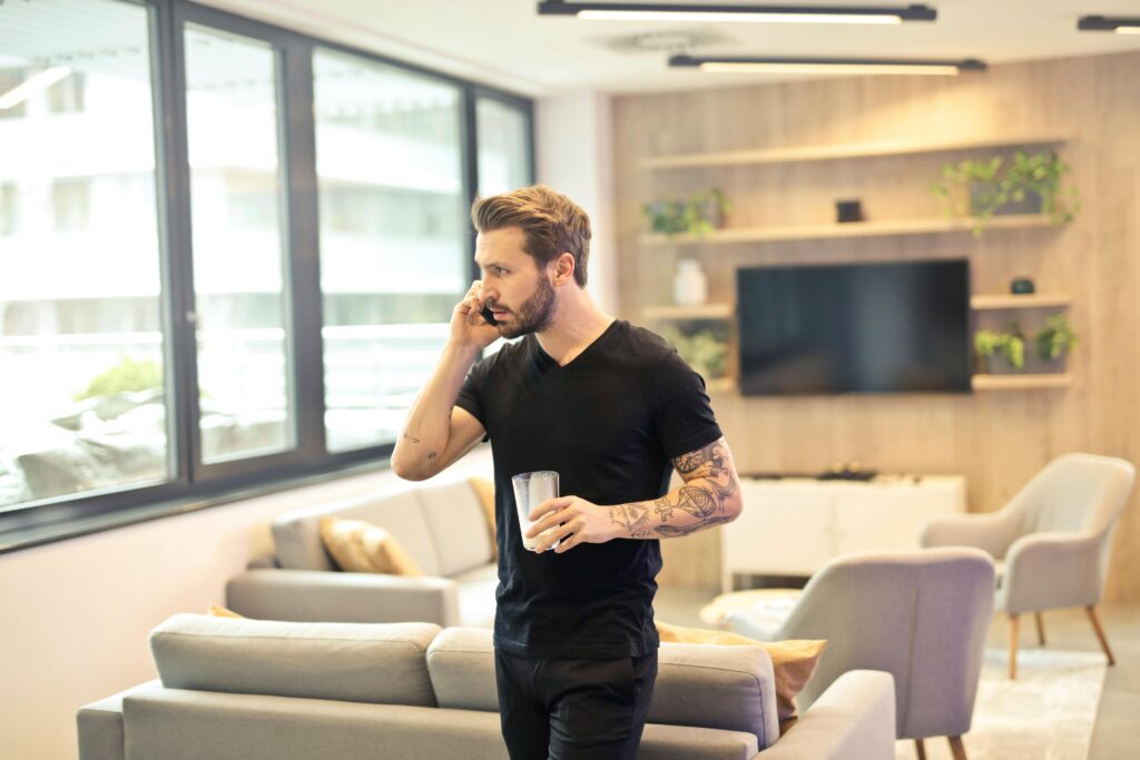 A tattooed man talking on a smartphone and holding a drink in a contemporary living room setting.
